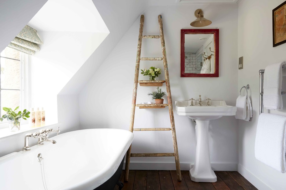 Modelo de cuarto de baño principal campestre pequeño con bañera exenta, paredes blancas, suelo de madera oscura, suelo marrón y lavabo con pedestal