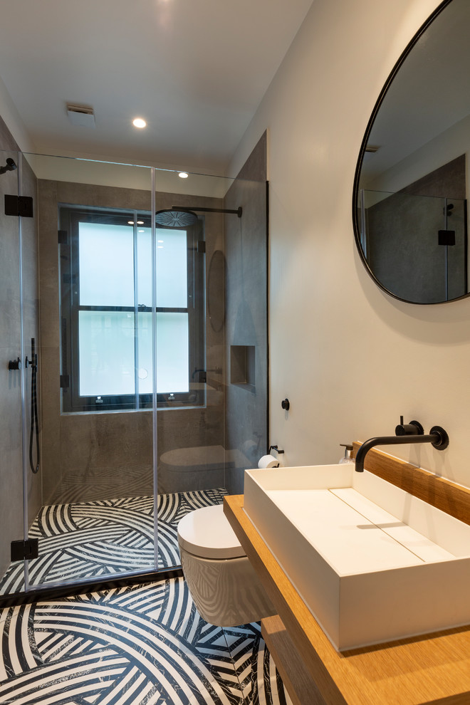 Cette image montre une salle de bain design de taille moyenne avec un placard sans porte, WC suspendus, un mur blanc, une vasque, un plan de toilette en bois, un sol multicolore, une cabine de douche à porte battante et un plan de toilette beige.