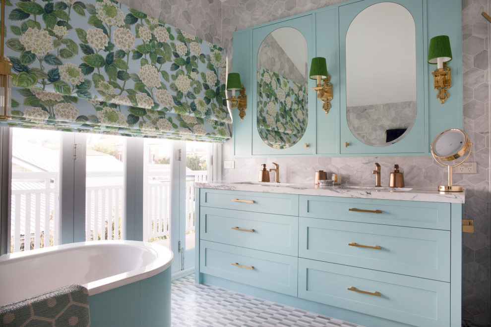 Aménagement d'une salle de bain bord de mer avec un placard à porte shaker, des portes de placard bleues, une baignoire indépendante, un carrelage gris, un lavabo encastré, un sol gris, un plan de toilette blanc, meuble double vasque et meuble-lavabo encastré.