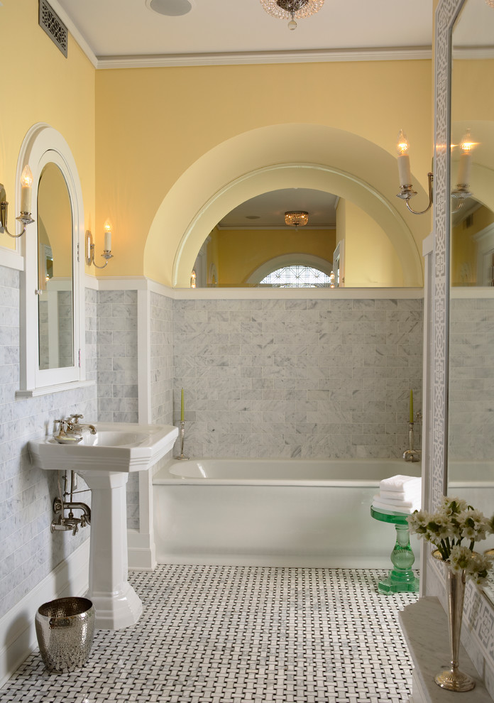 Klassisk inredning av ett en-suite badrum, med ett piedestal handfat, gula väggar, ett badkar i en alkov, grå kakel, stenkakel, marmorgolv och flerfärgat golv