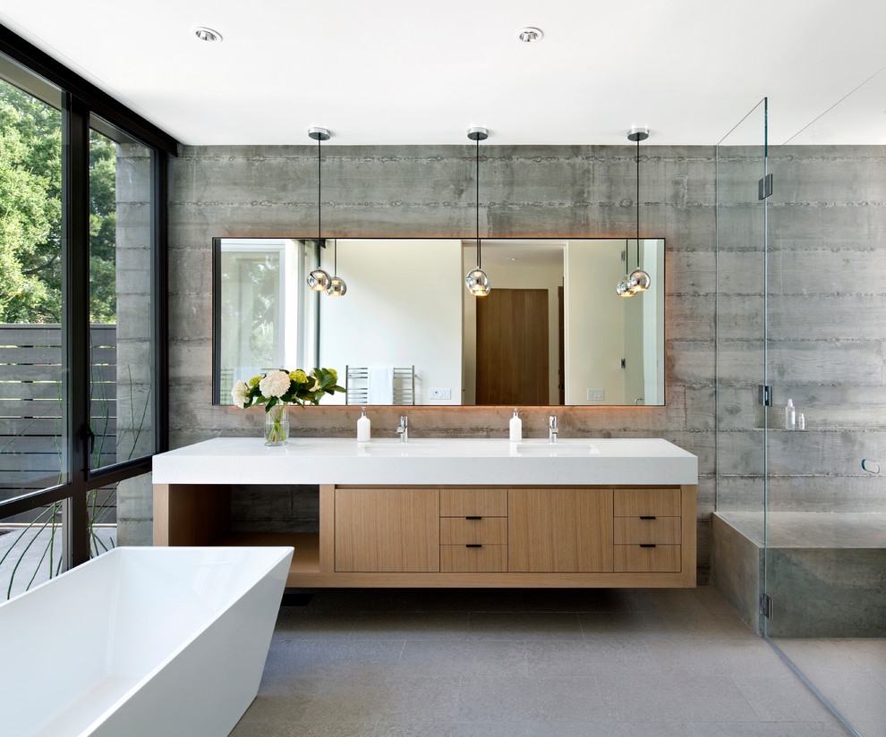 Modelo de cuarto de baño principal actual con armarios con paneles lisos, puertas de armario de madera clara, bañera exenta, ducha a ras de suelo, paredes grises y lavabo integrado