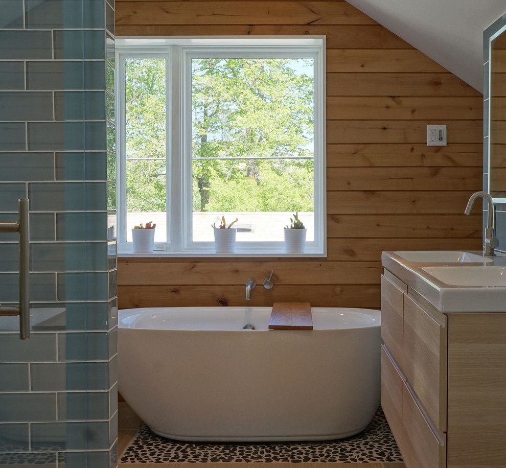 Photo of a medium sized scandinavian ensuite bathroom in Denver with flat-panel cabinets, light wood cabinets, a freestanding bath, a walk-in shower, a two-piece toilet, blue tiles, glass tiles, white walls, pebble tile flooring, an integrated sink and solid surface worktops.