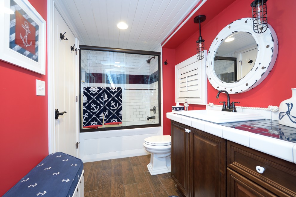 グランドラピッズにある中くらいなトランジショナルスタイルのおしゃれな浴室 (レイズドパネル扉のキャビネット、中間色木目調キャビネット、アルコーブ型浴槽、シャワー付き浴槽	、分離型トイレ、青いタイル、サブウェイタイル、赤い壁、セラミックタイルの床、オーバーカウンターシンク、タイルの洗面台) の写真