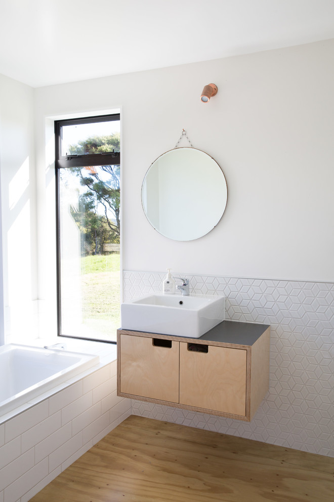 Inredning av ett nordiskt badrum med dusch, med släta luckor, skåp i ljust trä, ett platsbyggt badkar, vit kakel, vita väggar, plywoodgolv, ett fristående handfat och brunt golv