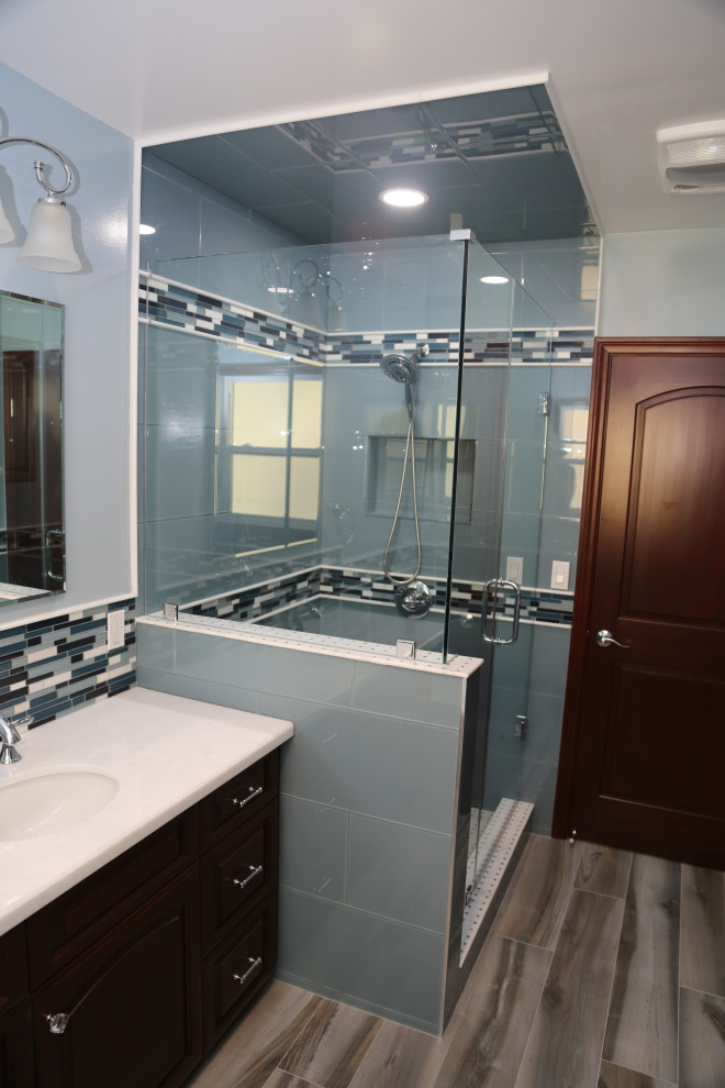 Cette image montre une douche en alcôve principale minimaliste en bois foncé de taille moyenne avec un placard avec porte à panneau surélevé, une baignoire en alcôve, WC à poser, un carrelage bleu, un carrelage en pâte de verre, un mur bleu, un sol en carrelage de porcelaine, un lavabo encastré, un plan de toilette en marbre, un sol bleu, une cabine de douche à porte battante, un plan de toilette blanc, un banc de douche, meuble simple vasque et meuble-lavabo encastré.