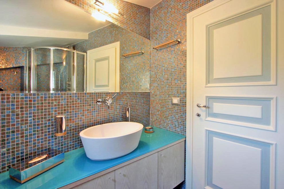 Modern inredning av ett stort badrum, med ett nedsänkt handfat, skåp i ljust trä, bänkskiva i glas, en hörndusch, en vägghängd toalettstol, blå kakel, mosaik, blå väggar, klinkergolv i keramik och släta luckor