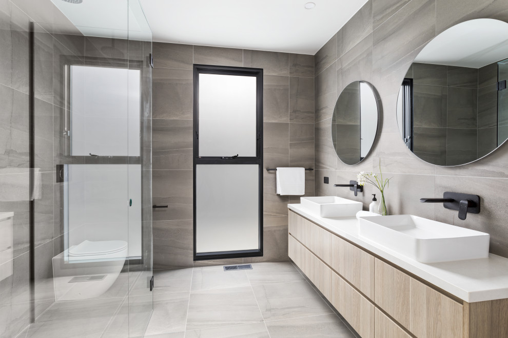 Modelo de cuarto de baño doble actual con armarios con paneles lisos, puertas de armario de madera clara, baldosas y/o azulejos grises, lavabo sobreencimera, suelo gris y encimeras blancas