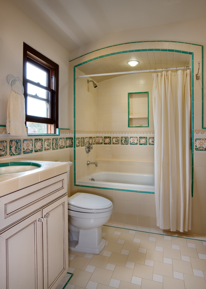 На фото: ванная комната в средиземноморском стиле с врезной раковиной, фасадами с выступающей филенкой, столешницей из плитки, душем над ванной, унитазом-моноблоком, разноцветной плиткой, керамической плиткой, желтыми стенами, полом из мозаичной плитки и бежевыми фасадами