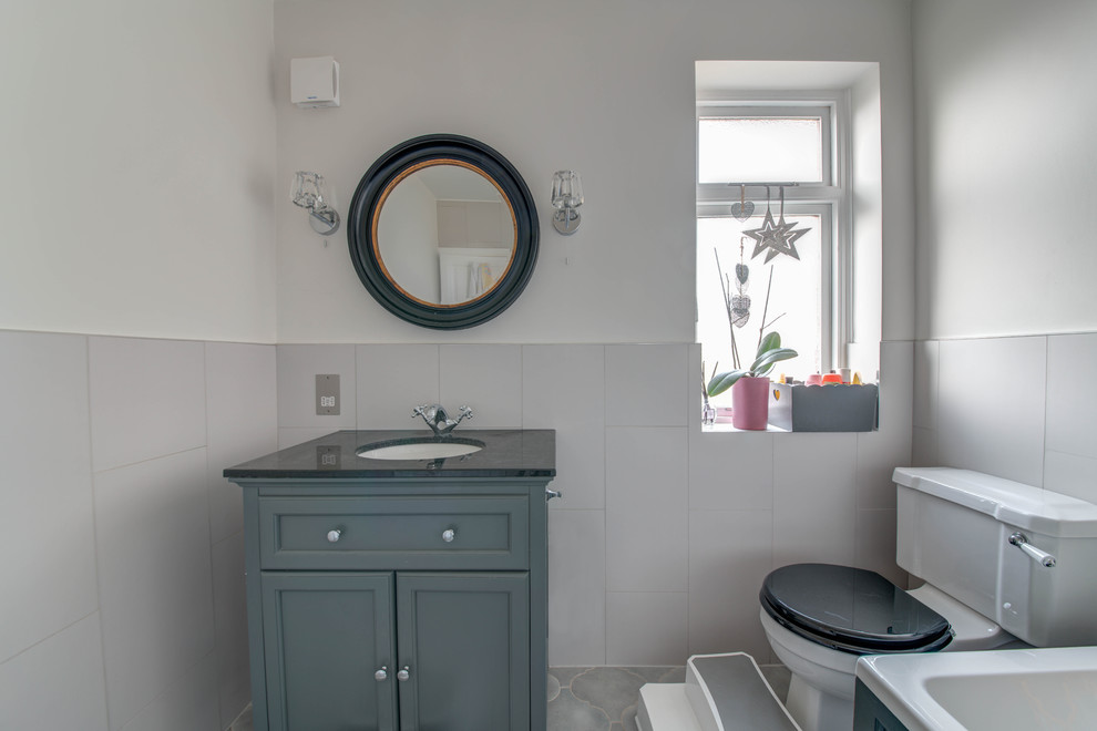 Photo of a medium sized classic family bathroom in London with shaker cabinets, grey cabinets, a built-in bath, a shower/bath combination, a two-piece toilet, grey tiles, ceramic tiles, grey walls, ceramic flooring, a built-in sink, marble worktops, grey floors, a hinged door and black worktops.