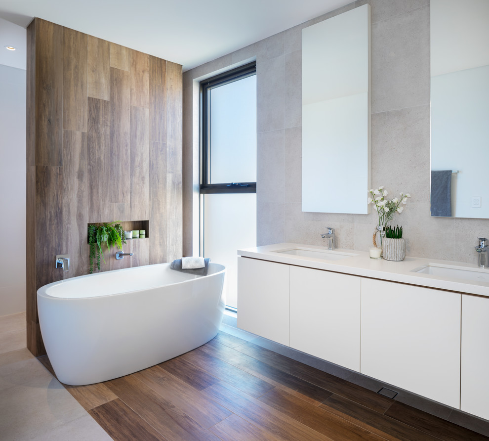 На фото: ванная комната в современном стиле с плоскими фасадами, белыми фасадами, отдельно стоящей ванной, серой плиткой, врезной раковиной, коричневым полом и белой столешницей с