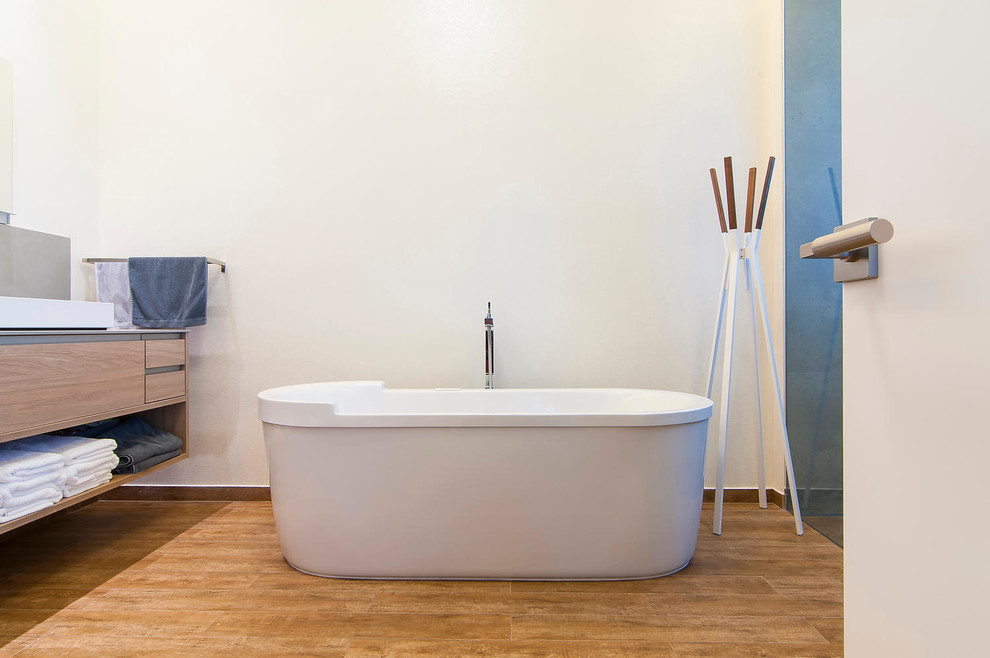 Foto på ett stort funkis en-suite badrum, med ett väggmonterat handfat, ett fristående badkar, en öppen dusch, en vägghängd toalettstol, släta luckor, skåp i ljust trä, kaklad bänkskiva, grå kakel, vita väggar och mellanmörkt trägolv