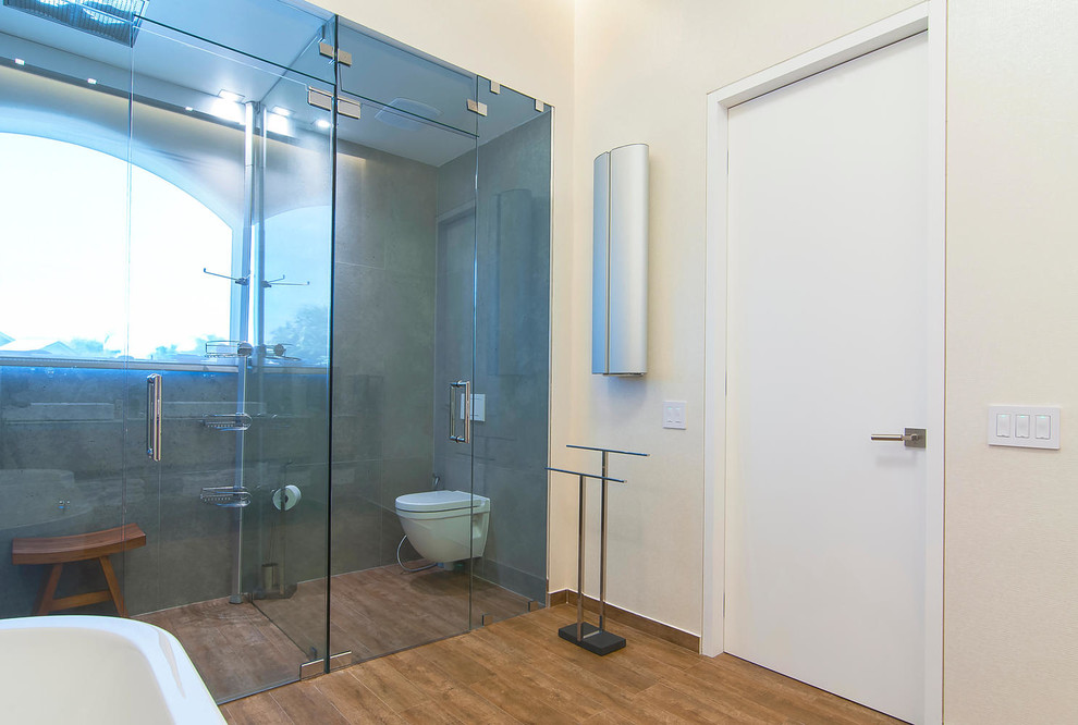 Réalisation d'une grande salle de bain principale design en bois clair avec un lavabo suspendu, une baignoire indépendante, une douche ouverte, WC suspendus, un placard à porte plane, un carrelage gris, un mur blanc et un sol en bois brun.