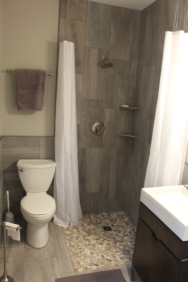 Modern inredning av ett litet badrum med dusch, med ett piedestal handfat, släta luckor, skåp i mörkt trä, en kantlös dusch, en toalettstol med separat cisternkåpa, grå kakel, kakel i småsten, grå väggar och kalkstensgolv