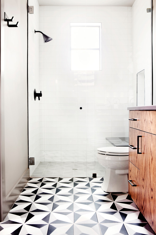 Klassisk inredning av ett litet badrum med dusch, med släta luckor, skåp i mellenmörkt trä, en dusch i en alkov, en toalettstol med separat cisternkåpa, vit kakel, porslinskakel, vita väggar, cementgolv, ett undermonterad handfat, bänkskiva i akrylsten, flerfärgat golv och dusch med gångjärnsdörr