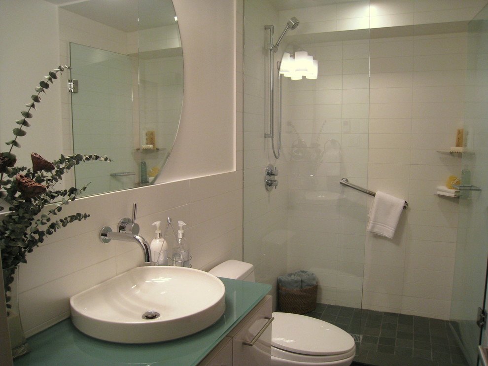 Exempel på ett litet modernt badrum med dusch, med en dusch i en alkov, en toalettstol med hel cisternkåpa, ett nedsänkt handfat, släta luckor, vita skåp, bänkskiva i glas, vit kakel, porslinskakel, vita väggar och skiffergolv