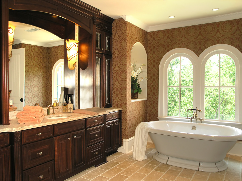 Foto på ett stort medelhavsstil en-suite badrum, med luckor med upphöjd panel, skåp i mörkt trä, ett fristående badkar, bruna väggar, kalkstensgolv, ett undermonterad handfat, granitbänkskiva och beiget golv
