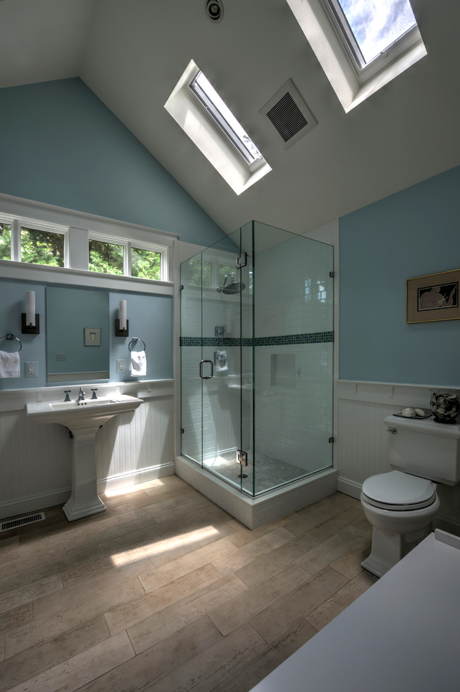 Bild på ett amerikanskt badrum med dusch, med en hörndusch, vit kakel, blå väggar, ljust trägolv och ett piedestal handfat