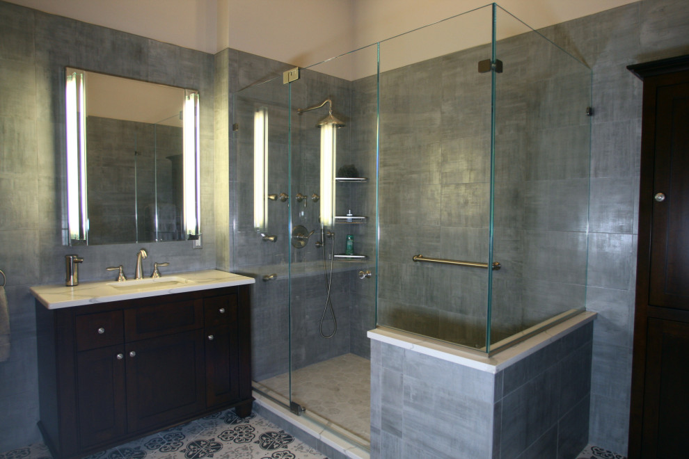 Example of a large transitional master gray tile and porcelain tile porcelain tile, multicolored floor, single-sink, vaulted ceiling and brick wall bathroom design in New York with brown cabinets, a one-piece toilet, gray walls, an undermount sink, quartz countertops, a hinged shower door, white countertops, a freestanding vanity and raised-panel cabinets