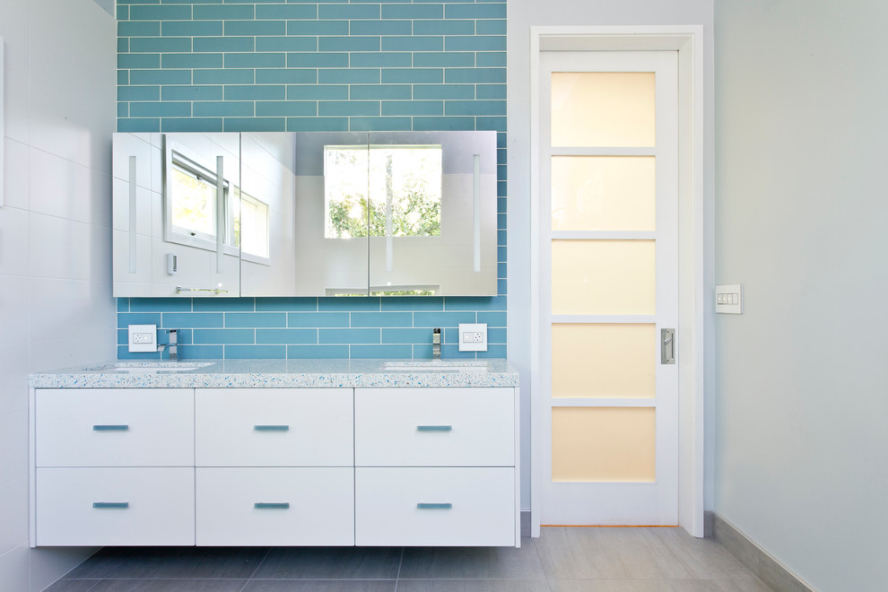 Пример оригинального дизайна: большая главная ванная комната в стиле модернизм с плоскими фасадами, белыми фасадами, синей плиткой, стеклянной плиткой, серыми стенами, полом из керамической плитки, врезной раковиной и столешницей из переработанного стекла
