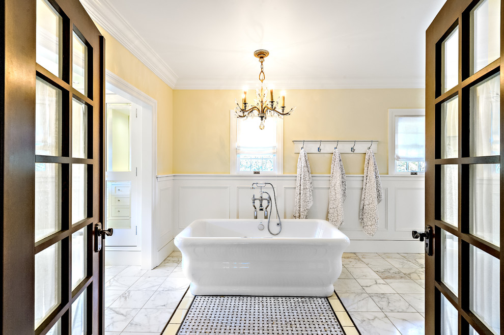 На фото: большая главная ванная комната в стиле неоклассика (современная классика) с фасадами с утопленной филенкой, белыми фасадами, отдельно стоящей ванной, серой плиткой, белой плиткой, каменной плиткой, желтыми стенами, мраморным полом, подвесной раковиной и душем в нише с