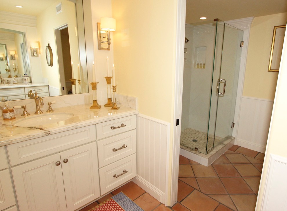 サンディエゴにある中くらいなトラディショナルスタイルのおしゃれなマスターバスルーム (レイズドパネル扉のキャビネット、白いキャビネット、コーナー設置型シャワー、白いタイル、セラミックタイル、黄色い壁、テラコッタタイルの床、アンダーカウンター洗面器、大理石の洗面台、置き型浴槽) の写真