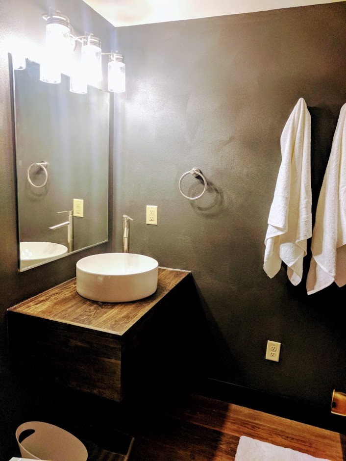 Idée de décoration pour une salle de bain chalet en bois vieilli de taille moyenne avec un placard à porte plane, un mur noir, un sol en bois brun, une vasque, un plan de toilette en bois et un sol marron.