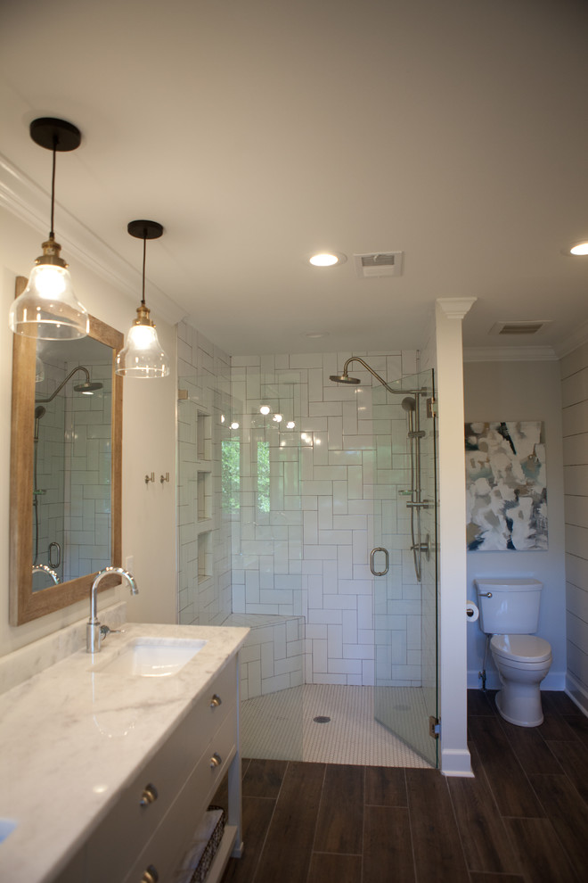 Modern inredning av ett stort en-suite badrum, med vita skåp, en hörndusch, en toalettstol med hel cisternkåpa, vit kakel, tunnelbanekakel, mörkt trägolv, marmorbänkskiva, grå väggar, brunt golv, dusch med gångjärnsdörr, ett undermonterad handfat och släta luckor