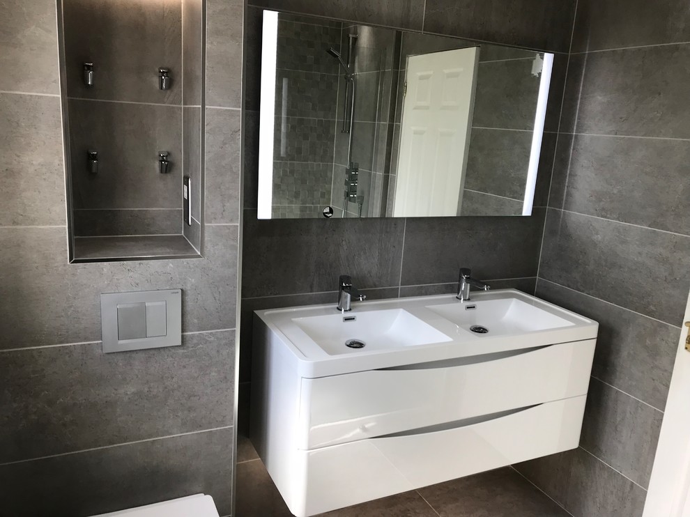 Modern inredning av ett mellanstort badrum för barn, med möbel-liknande, vita skåp, en öppen dusch, en vägghängd toalettstol, grå kakel, keramikplattor, grå väggar, klinkergolv i keramik, ett väggmonterat handfat, grått golv och med dusch som är öppen