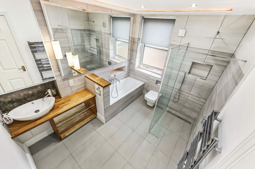 Inspiration for a large modern bathroom in London with glass-front cabinets, grey cabinets, a built-in bath, a wall mounted toilet, grey tiles, ceramic tiles, grey walls, porcelain flooring, a vessel sink, wooden worktops, grey floors, an open shower and brown worktops.