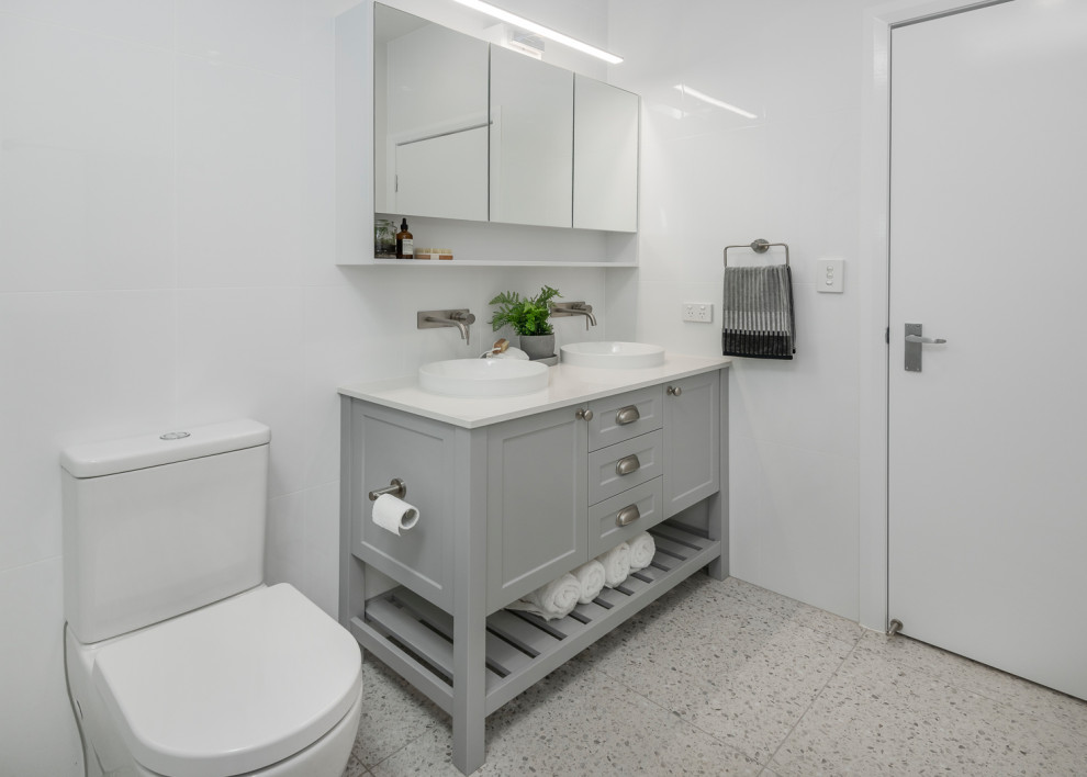 На фото: ванная комната среднего размера в стиле неоклассика (современная классика) с фасадами в стиле шейкер, серыми фасадами, открытым душем, унитазом-моноблоком, белой плиткой, керамогранитной плиткой, белыми стенами, полом из терраццо, душевой кабиной, настольной раковиной, столешницей из искусственного камня, серым полом, открытым душем и белой столешницей с
