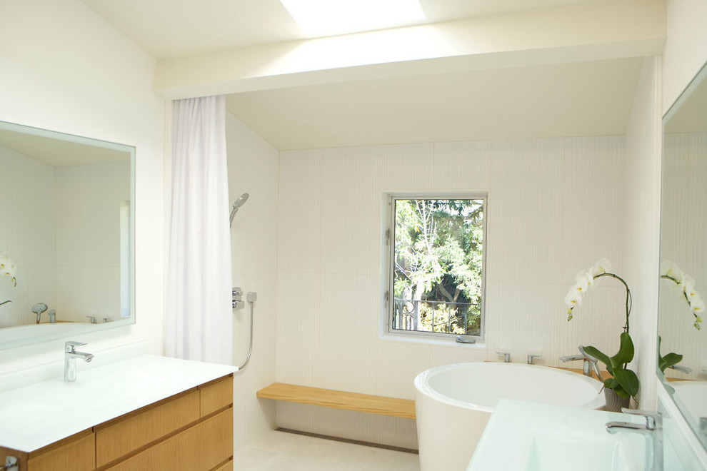 На фото: большая главная ванная комната в современном стиле с плоскими фасадами, светлыми деревянными фасадами, японской ванной, душем без бортиков, унитазом-моноблоком, белой плиткой, керамической плиткой, белыми стенами, полом из керамической плитки, монолитной раковиной, столешницей из переработанного стекла, белым полом, шторкой для ванной и белой столешницей