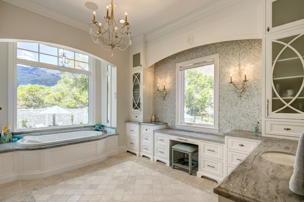 Design ideas for a large classic ensuite bathroom in Salt Lake City with recessed-panel cabinets, white cabinets, a built-in bath, grey tiles, glass tiles, beige walls, travertine flooring, a submerged sink, granite worktops, beige floors and grey worktops.