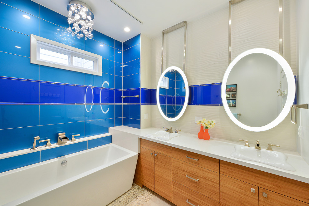 На фото: главная ванная комната среднего размера в стиле модернизм с плоскими фасадами, светлыми деревянными фасадами, отдельно стоящей ванной, душем над ванной, синей плиткой, стеклянной плиткой, белыми стенами, полом из галечной плитки, монолитной раковиной и столешницей из искусственного камня с