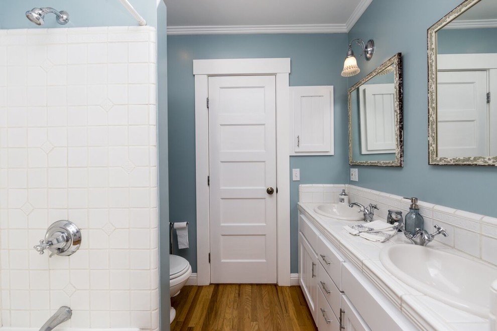 Modern inredning av ett badrum, med släta luckor, vita skåp, en dusch/badkar-kombination, vit kakel, keramikplattor, blå väggar, mellanmörkt trägolv, ett nedsänkt handfat och kaklad bänkskiva
