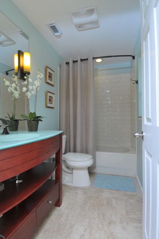 Foto på ett funkis badrum, med möbel-liknande, skåp i mellenmörkt trä, bänkskiva i glas, ett badkar i en alkov, en dusch/badkar-kombination, en toalettstol med separat cisternkåpa, vit kakel och tunnelbanekakel