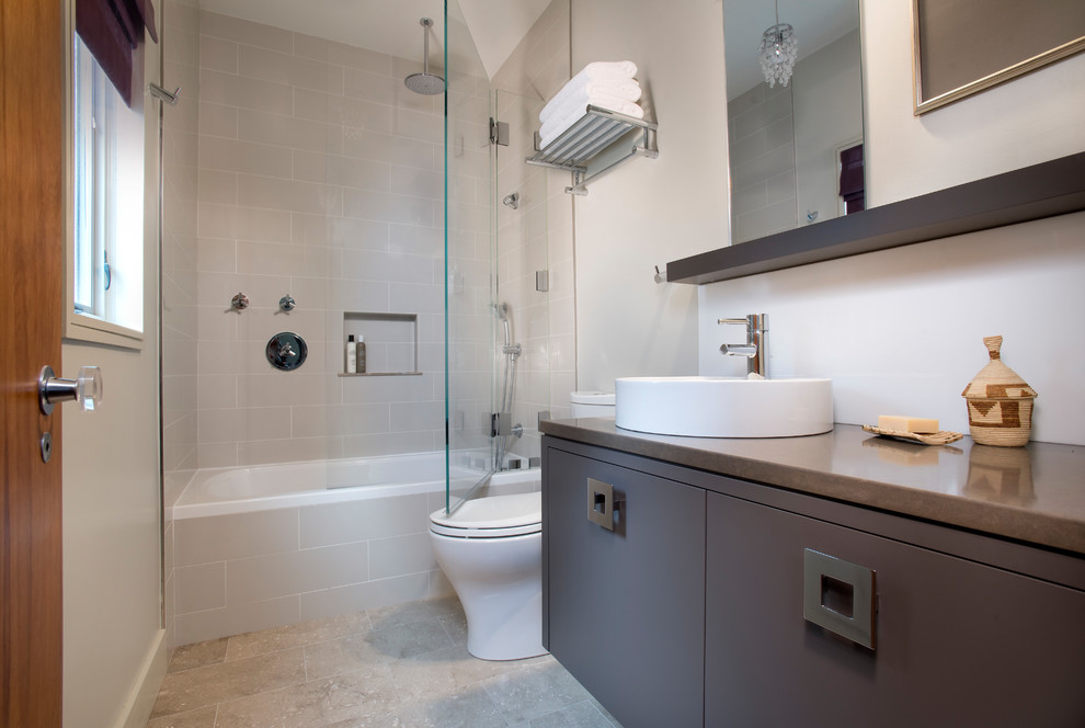 Bild på ett litet funkis badrum, med släta luckor, bruna skåp, ett badkar i en alkov, en dusch/badkar-kombination, en toalettstol med separat cisternkåpa, grå kakel, keramikplattor, grå väggar, kalkstensgolv, ett fristående handfat, bänkskiva i kvarts, grått golv och dusch med gångjärnsdörr