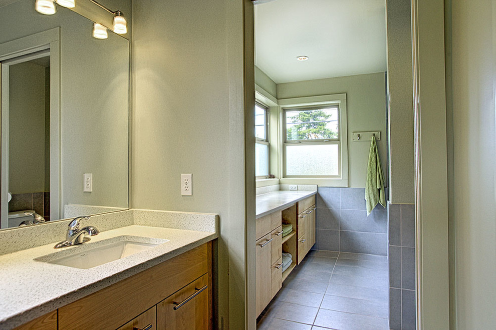 Inredning av ett modernt badrum, med ett undermonterad handfat, släta luckor, skåp i ljust trä, bänkskiva i kvarts, ett badkar i en alkov, en dusch/badkar-kombination, en toalettstol med separat cisternkåpa, grå kakel, keramikplattor, gröna väggar och klinkergolv i keramik