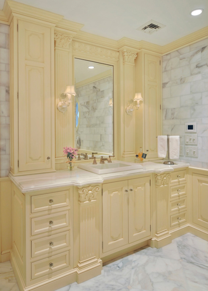 Photo of a large classic grey and yellow ensuite bathroom in New York with a built-in sink, yellow cabinets, marble flooring, a walk-in shower, a two-piece toilet, white tiles, stone tiles, white walls, marble worktops and raised-panel cabinets.