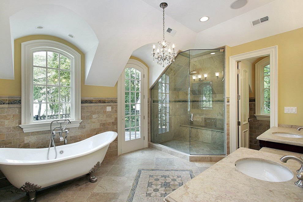 Идея дизайна: огромная главная ванная комната в стиле неоклассика (современная классика) с ванной на ножках, душем в нише, бежевой плиткой, коричневой плиткой, серой плиткой, керамогранитной плиткой, желтыми стенами, полом из керамогранита, врезной раковиной, столешницей из известняка, бежевым полом, душем с распашными дверями и бежевой столешницей