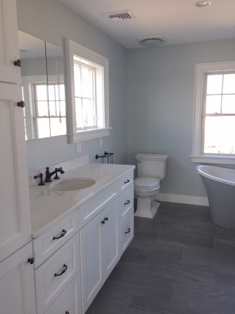 Cette photo montre une grande salle de bain principale bord de mer avec un placard avec porte à panneau encastré, une baignoire indépendante, WC à poser, un sol en carrelage de céramique, un lavabo intégré, un plan de toilette en marbre, un sol gris, des portes de placard blanches et un mur bleu.