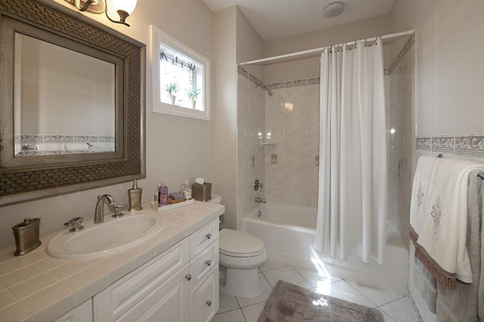 This is an example of a large classic ensuite bathroom in DC Metro with a built-in sink, raised-panel cabinets, white cabinets, tiled worktops, an alcove bath, a shower/bath combination, a two-piece toilet, white tiles, ceramic tiles, white walls, ceramic flooring, white floors and a shower curtain.