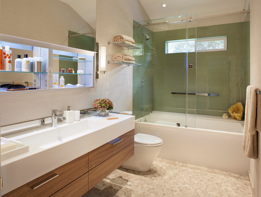 This is an example of a medium sized contemporary ensuite bathroom in San Francisco with flat-panel cabinets, medium wood cabinets, an alcove bath, a shower/bath combination, a two-piece toilet, green tiles, glass tiles, beige walls, pebble tile flooring, an integrated sink and solid surface worktops.
