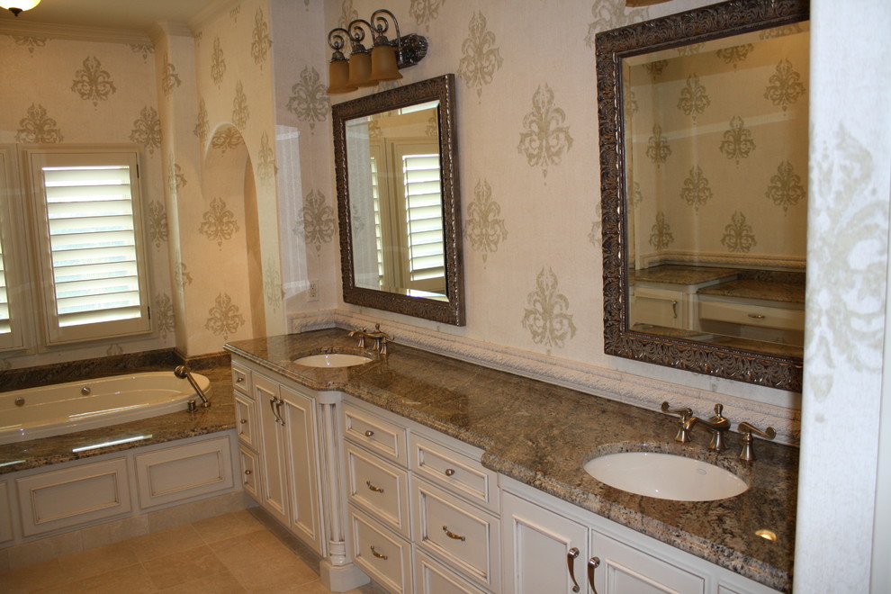Example of a large classic master beige tile and stone tile travertine floor drop-in bathtub design in Other with an undermount sink, flat-panel cabinets, distressed cabinets, granite countertops and beige walls