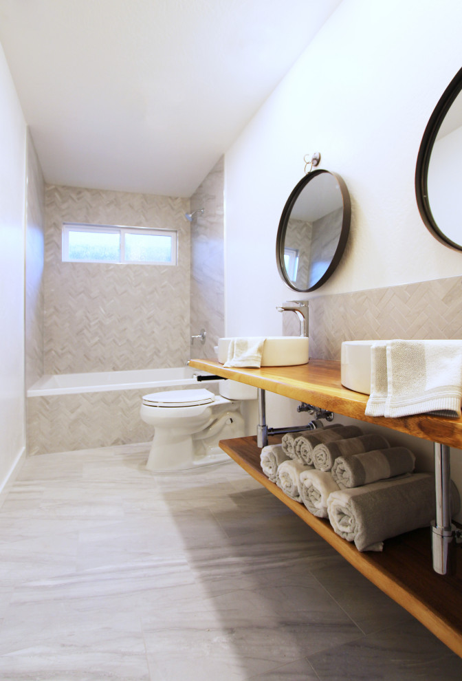 Inredning av ett litet brun brunt badrum för barn, med öppna hyllor, skåp i mellenmörkt trä, ett badkar i en alkov, en dusch/badkar-kombination, en toalettstol med separat cisternkåpa, grå kakel, porslinskakel, vita väggar, klinkergolv i porslin, ett fristående handfat, träbänkskiva och grått golv