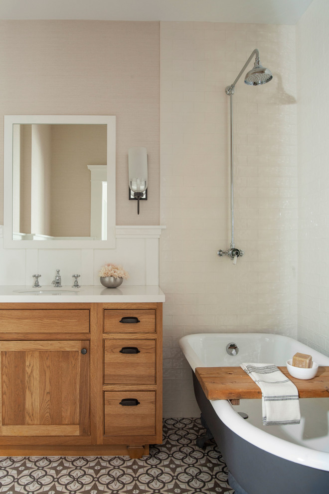 Inredning av ett klassiskt badrum, med luckor med profilerade fronter, skåp i mellenmörkt trä, ett badkar med tassar, beige kakel, tunnelbanekakel, beige väggar, ett undermonterad handfat och flerfärgat golv