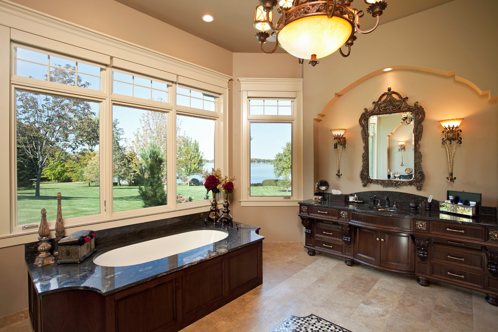 На фото: ванная комната в классическом стиле с врезной раковиной, темными деревянными фасадами, полновстраиваемой ванной, бежевой плиткой, окном и фасадами с утопленной филенкой с
