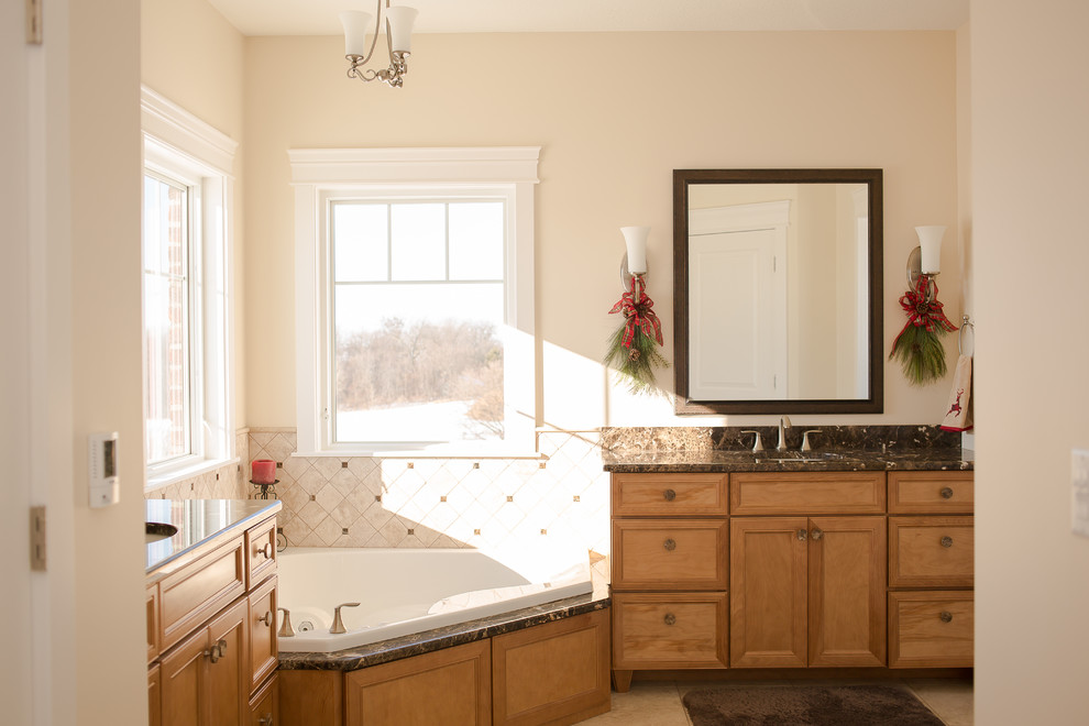 Cette image montre une salle de bain traditionnelle en bois clair avec un lavabo encastré, un placard à porte plane, un plan de toilette en granite, une baignoire d'angle, un carrelage beige et des carreaux de céramique.