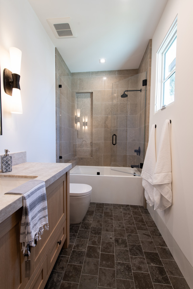 Mid-sized transitional gray tile and marble tile vinyl floor, gray floor and single-sink alcove shower photo in Orange County with recessed-panel cabinets, light wood cabinets, a one-piece toilet, white walls, an undermount sink, limestone countertops, a hinged shower door, beige countertops and a floating vanity