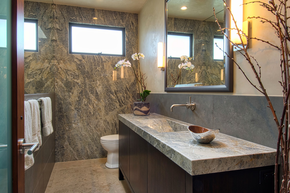 Réalisation d'une salle de bain design en bois foncé avec un placard à porte plane, un carrelage gris, des dalles de pierre, un mur beige, un lavabo intégré, un sol beige, un plan de toilette en granite, une fenêtre et un mur en pierre.