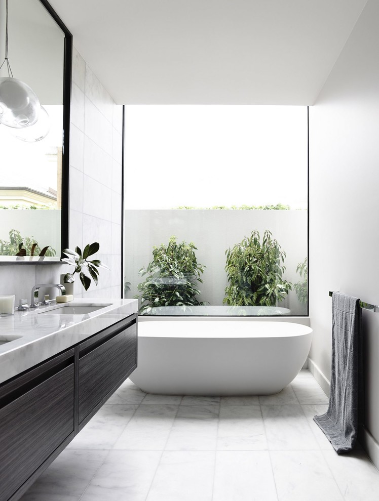 Imagen de cuarto de baño contemporáneo con armarios con paneles lisos, puertas de armario grises, bañera exenta, baldosas y/o azulejos blancos, paredes blancas, lavabo bajoencimera y suelo blanco